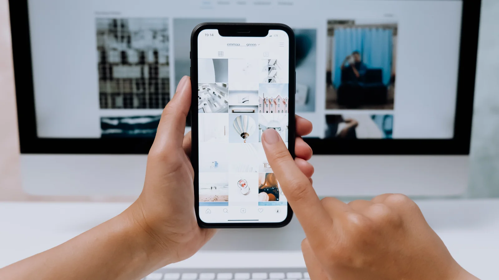 Hands browsing an Instagram photo grid on a smartphone in front of a computer